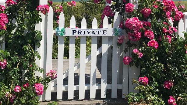 A white gate with a 'PRIVATE' sign, surrounded by pink flowers and a clear view of the ocean. - In-Town • Walk to Beaches • Charming Cottage • Private Patio & Easy Parking (Nantucket)
