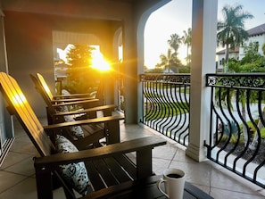 A wooden bench with a patterned cushion, a white mug, and a view of a gated balcony with palm trees and a house in the background. - Spacious Waterfront Naples Pool Home-Minutes to Ritz-Carlton & Vanderbilt Beach (Naples)