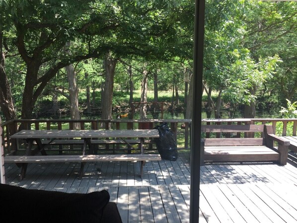 An outdoor deck with wooden benches and picnic tables, surrounded by trees and a body of water.