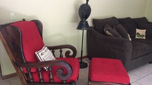 A wooden chair with a red cushion and a matching ottoman, a floor lamp, and a dark-colored sofa with cushions. - STUNNING Private Frio River! Ping Pong, Guest Cabin, Rope Swing, Kayak Heaven (Leakey)