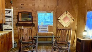 A rustic wooden cabin interior with two wooden chairs, a small table, a window with a view, a heater, and a lantern. - STUNNING Private Frio River! Ping Pong, Guest Cabin, Rope Swing, Kayak Heaven (Leakey)