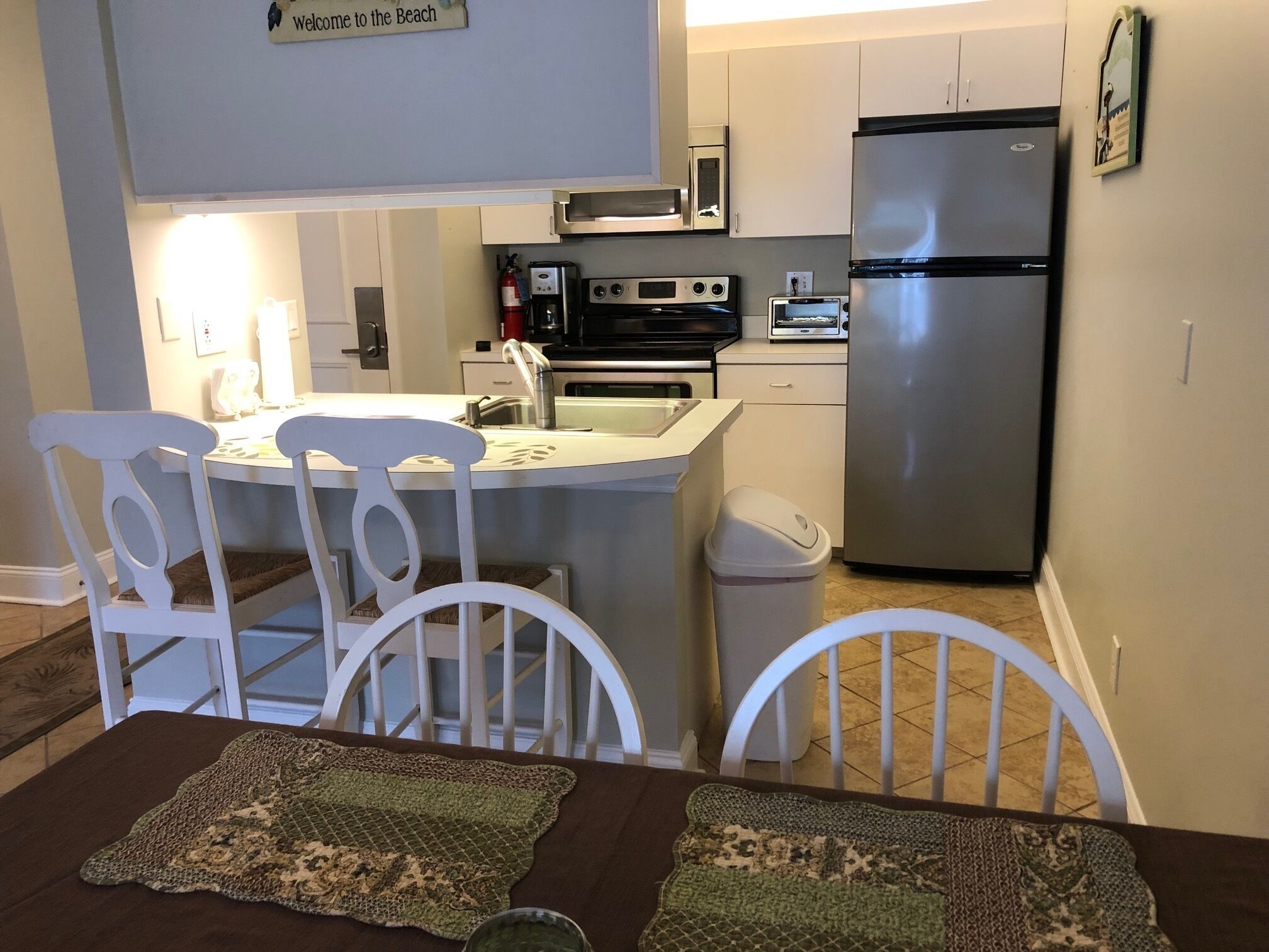 A compact kitchen with a refrigerator, microwave, and oven. There is a small trash bin and a sign that says 'Welcome to the Beach.'