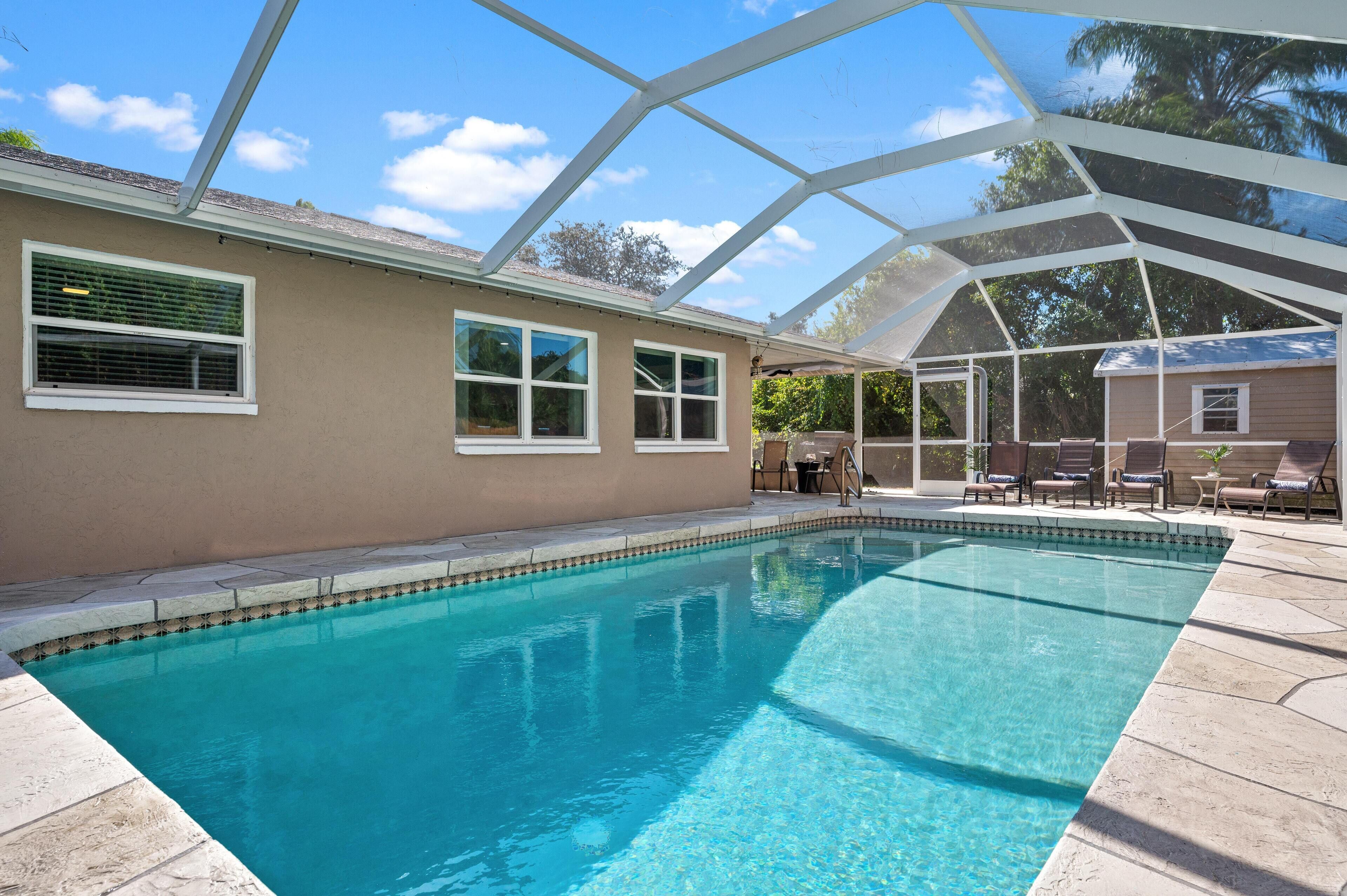 A clear swimming pool with a covered patio area, a beige building with windows, and a clear blue sky.