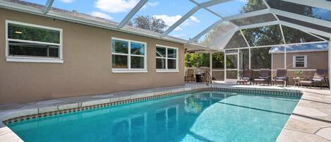 A clear swimming pool with a covered patio area, a beige building with windows, and a clear blue sky.