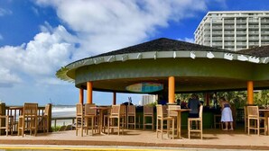 A covered outdoor seating area with wooden chairs and tables, a view of a bridge, and a building in the background. - Royale Palms 14022H Oceanview Studio in Kingston (Myrtle Beach)