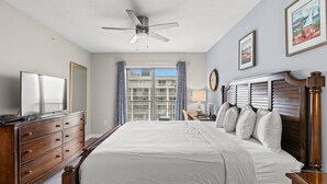 A bedroom with a large bed, wooden dresser, ceiling fan, and a view of a building through the window. - Royale Palms 14022H Oceanview Studio in Kingston (Myrtle Beach)