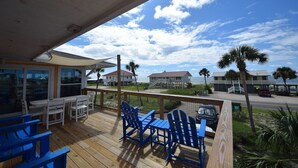 A wooden deck with blue Adirondack chairs and a table, overlooking a beach with houses and palm trees. - Summer Sale! Steps to Beach, Amazing View! Pool, Game room, Hot-tub! (St George Island)