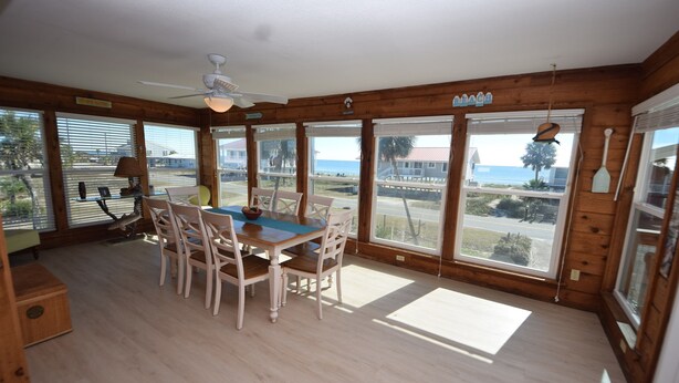A wooden-paneled room with a dining table and chairs, large windows with views of the ocean, and a ceiling fan. - Summer Sale! Steps to Beach, Amazing View! Pool, Game room, Hot-tub! (St George Island)