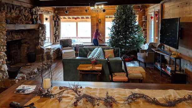 L’intérieur d’un chalet confortable, avec des murs en rondins de bois, une cheminée en pierre, un canapé vert, un sapin de Noël et un téléviseur fixé au mur.