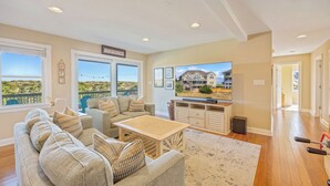 A spacious living room with a large window, a sofa set, a coffee table, and a television. - Premier Oceanfront Dog Friendly Home w/ Pool, Hot Tub, EV Charger, and Game Room (Salvo)