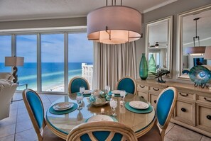Un comedor con vista al océano, una mesa redonda dispuesta con platos y vasos, y una lámpara colgante grande arriba.