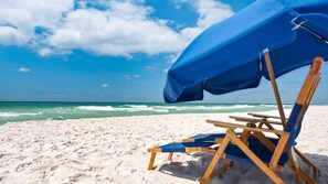 Una silla de playa bajo una sombrilla azul en una playa arenosa, con el océano al fondo.