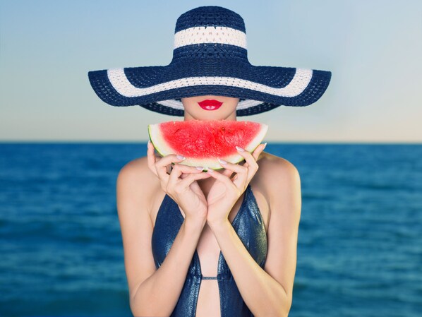 Una persona sostiene una tajada de sandía, vestida con un bañador azul y un sombrero de ala ancha, con una masa de agua al fondo.