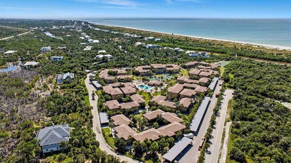 An aerial view of a resort with a central pool, surrounded by residential buildings, a beach, and a river.