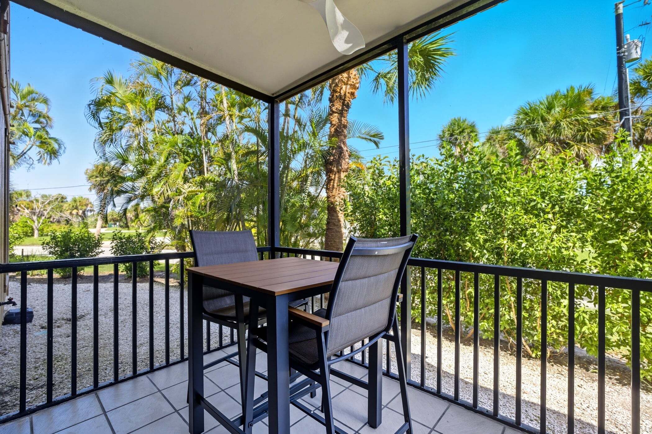 A covered balcony with a table and chairs, surrounded by a metal railing, overlooking a garden with palm trees and other greenery.