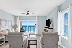 A living room with a sofa, chairs, a TV, and a balcony with a view of the ocean.