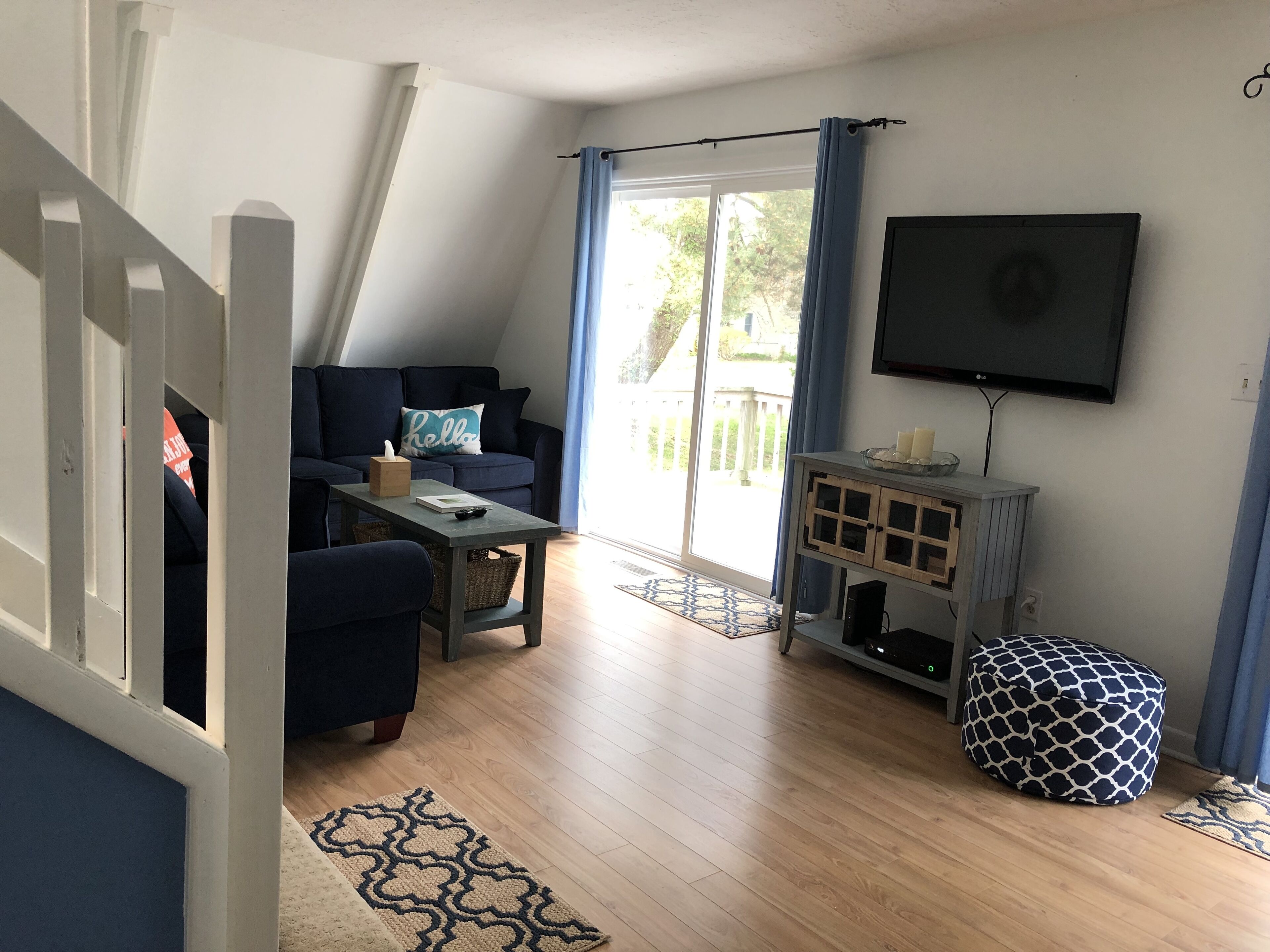 Sala de estar con un sofá azul oscuro, una mesita con un control remoto, una cesta y un tapete estampado. Hay un televisor montado en la pared y una ventana con cortinas.