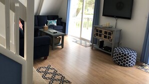 Un salon avec un canapé bleu foncé, une petite table avec une télécommande, un panier et un tapis à motifs. Un téléviseur est fixé au mur et il y a une fenêtre avec des rideaux.