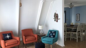 Un salon avec deux fauteuils orange, un fauteuil turquoise, un petit pouf et un coin repas comprenant une table et des chaises.