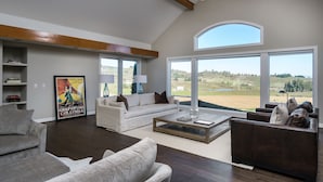 A spacious living room with a high ceiling, large windows offering a view of the countryside, and a variety of seating options including sofas and a coffee table.