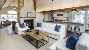 A spacious living room with a fireplace, white sofas with blue cushions, a wooden coffee table, and a kitchen area with bar stools.
