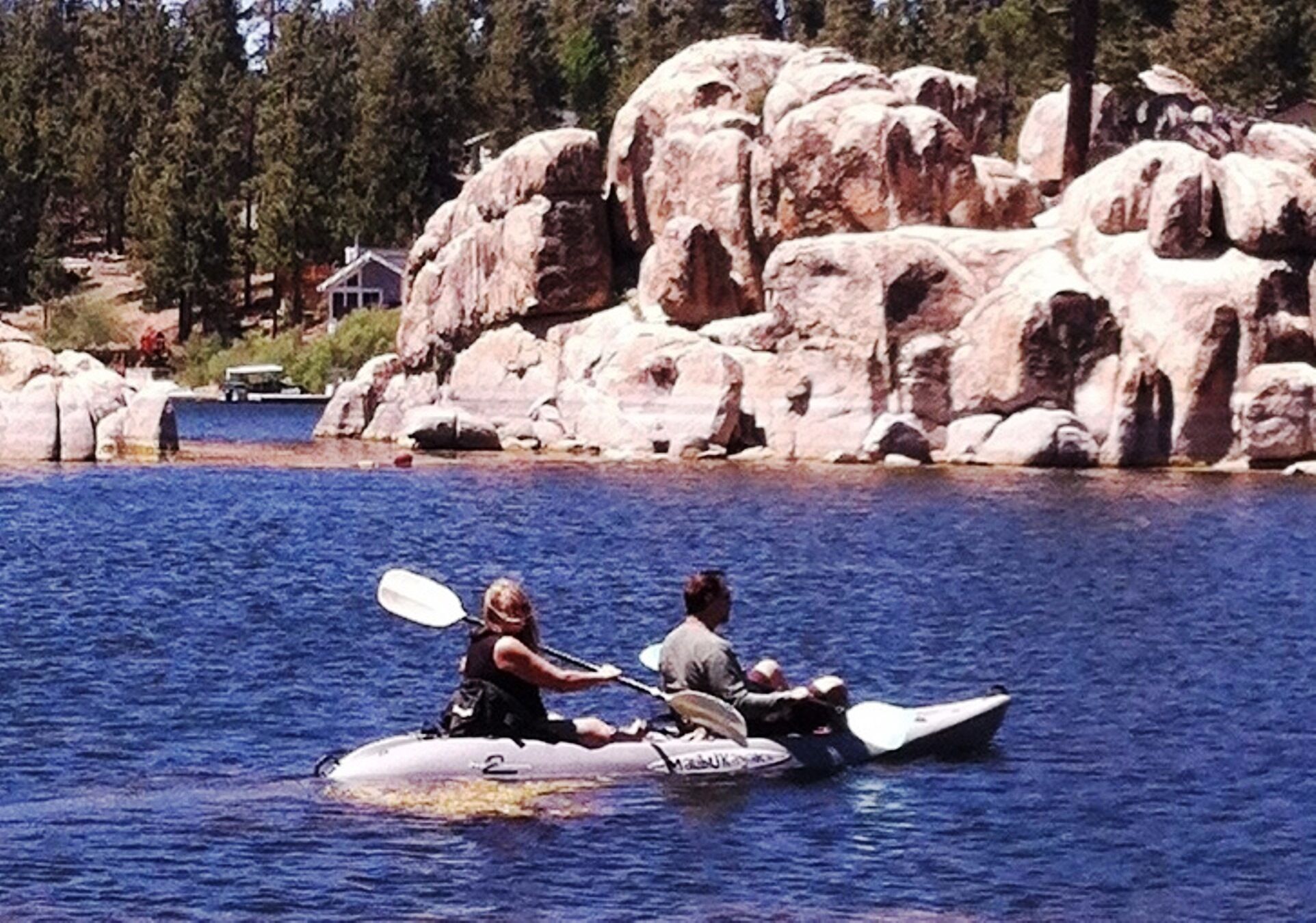 Dos personas kayakendo en un lago tranquilo con grandes rocas y una casa al fondo.