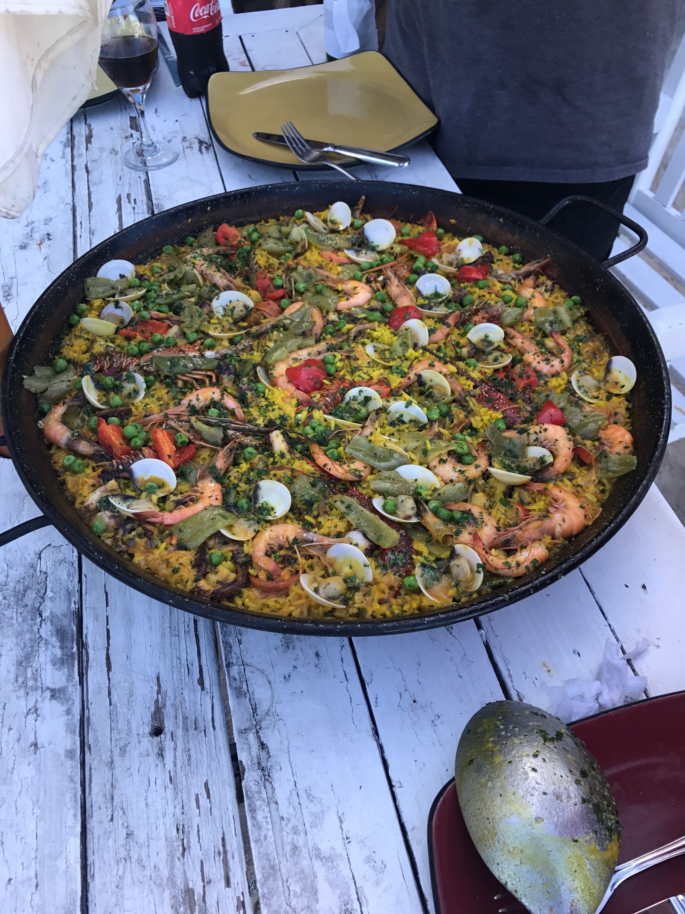 Una paella de mariscos grande sobre una mesa de madera blanca, con un tenedor y una cuchara al lado.