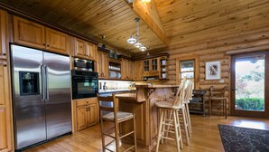 Cocina de cabaña de madera con refrigerador de acero inoxidable, armarios de madera, una zona de bar con taburetes y un amplio ventanal con vistas al exterior.