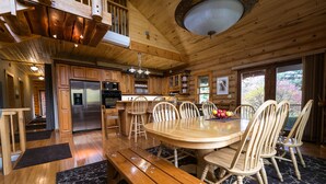 Interior de una cabaña de madera con comedor, cocina y ventanas amplias.