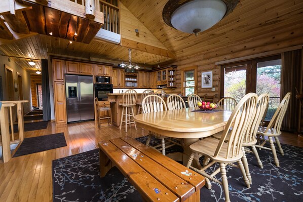 A wooden cabin interior with a dining area, kitchen, and large windows.