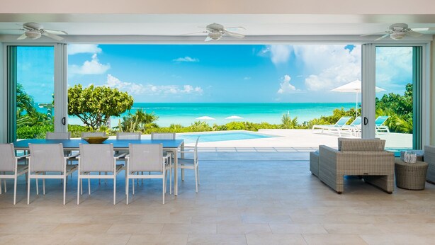 A spacious room with a large glass door leading to a pool and ocean view. There are white chairs and a dining table inside. - Villa Blue Heaven-Just Steps to  Beach and Turquoise Water -  Best Sunsets!  (Providenciales)