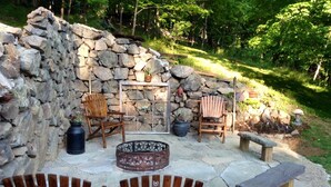 An outdoor patio with stone walls, two wooden Adirondack chairs, a fire pit, and a stone bench.