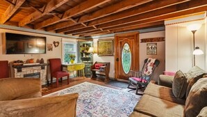A cozy living room with a stone fireplace, wooden beams, a red armchair, a patterned rug, and a wooden door with a decorative glass panel.