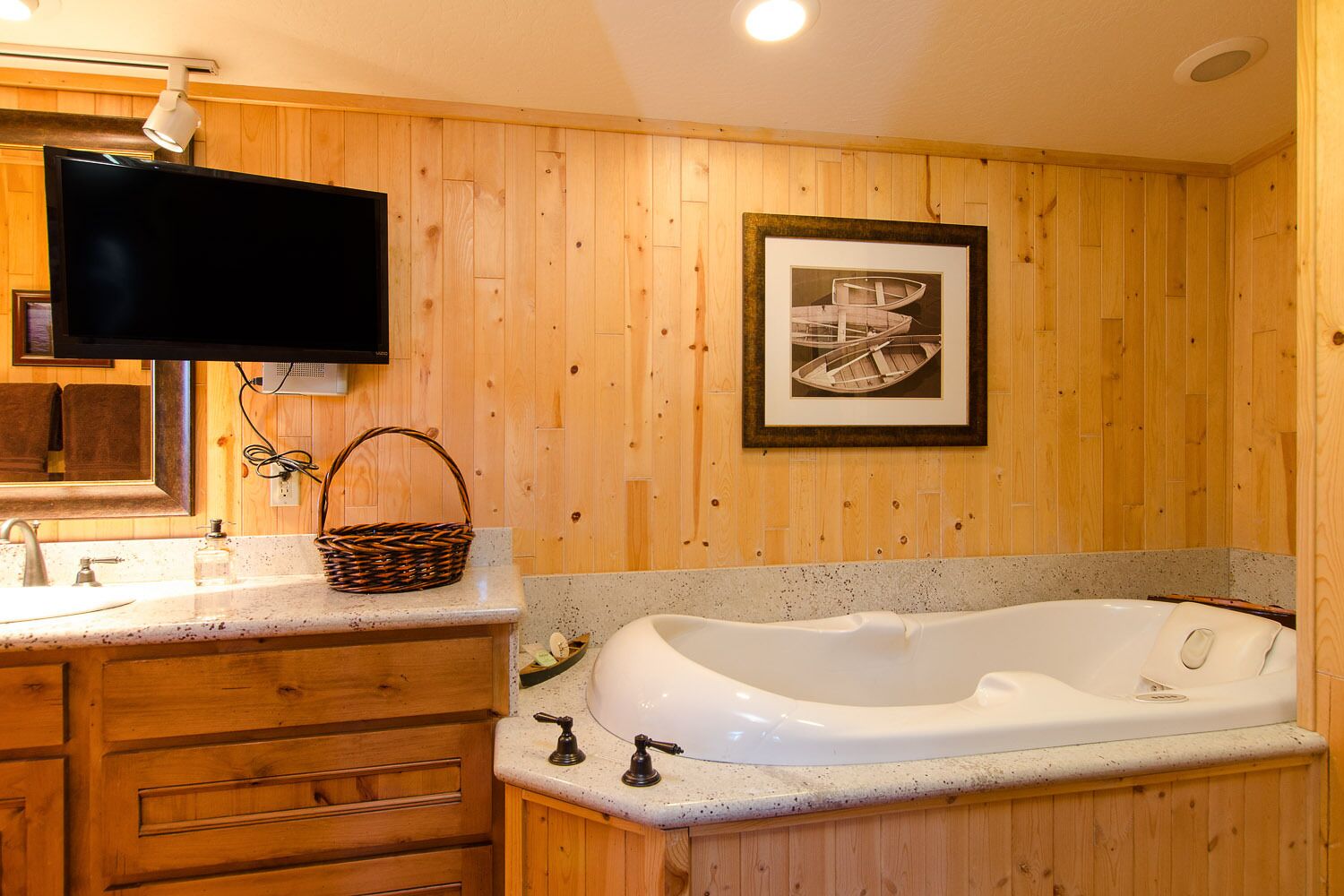Ein Badezimmer mit Holzpaneelen, einer großen Badewanne, einem Waschbecken mit Spiegel, einem darüber montierten Fernseher und einem gerahmten Bild an der Wand.