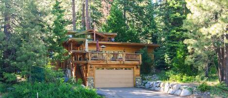 A two-story log cabin with a balcony, surrounded by dense forest.