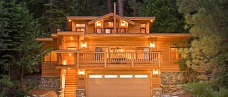 A large, two-story wooden cabin with a balcony, surrounded by trees.