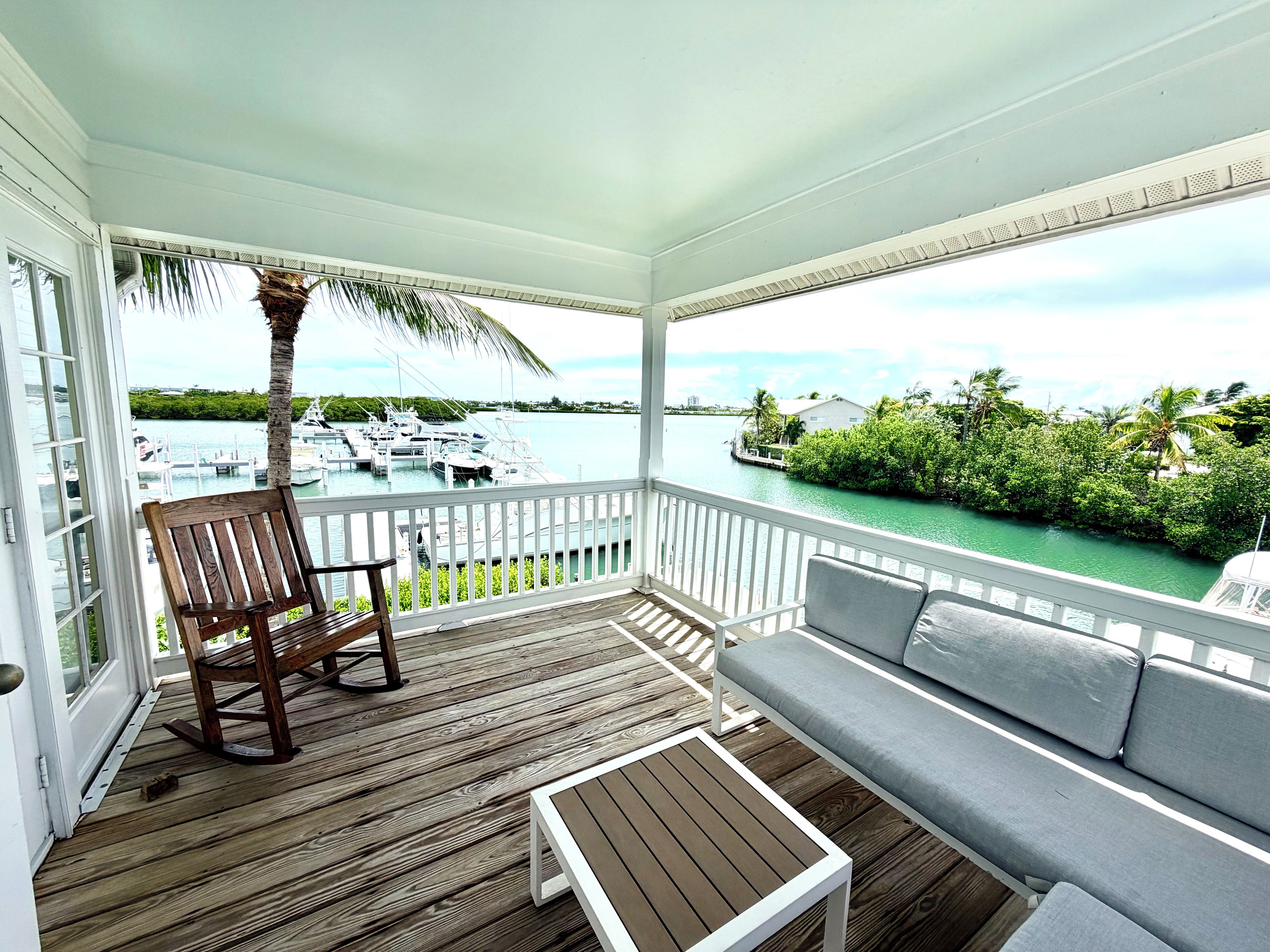 A covered porch with a wooden floor, a rocking chair, a gray sofa, and a view of a marina with boats and greenery.