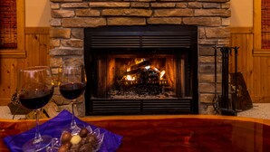 A stone fireplace with a fire, two glasses of red wine, and a bowl of snacks on a wooden table. - Charming 3BR / 3BA With Hot Tub, Fireplace & Pool Table (Galena)