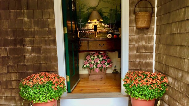 Une porte en bois, avec deux plantes en pot et des citrouilles disposées sur un paillasson à rayures.