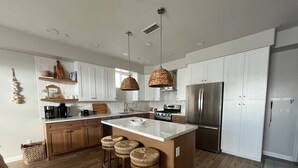 Una cocina moderna con armarios blancos, un refrigerador de acero inoxidable, una isla central con taburetes y una lámpara colgante de fibras naturales.