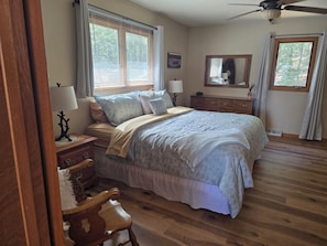 Un dormitorio con cama, mesitas de noche, una silla, cómoda, ventilador de techo y ventana con persianas.