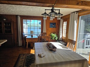 Ein Holzhütten-Interieur mit Esstisch, Stühlen, einer Lampe, einem Blumentopf und Blick auf einen See durch große Fenster.