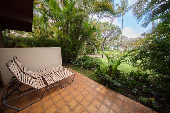 A patio with a wooden lounge chair, surrounded by lush greenery and a view of a garden.