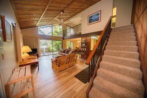 A spacious living area with a wooden staircase, a balcony with a view of palm trees, and a dining area with wicker furniture.