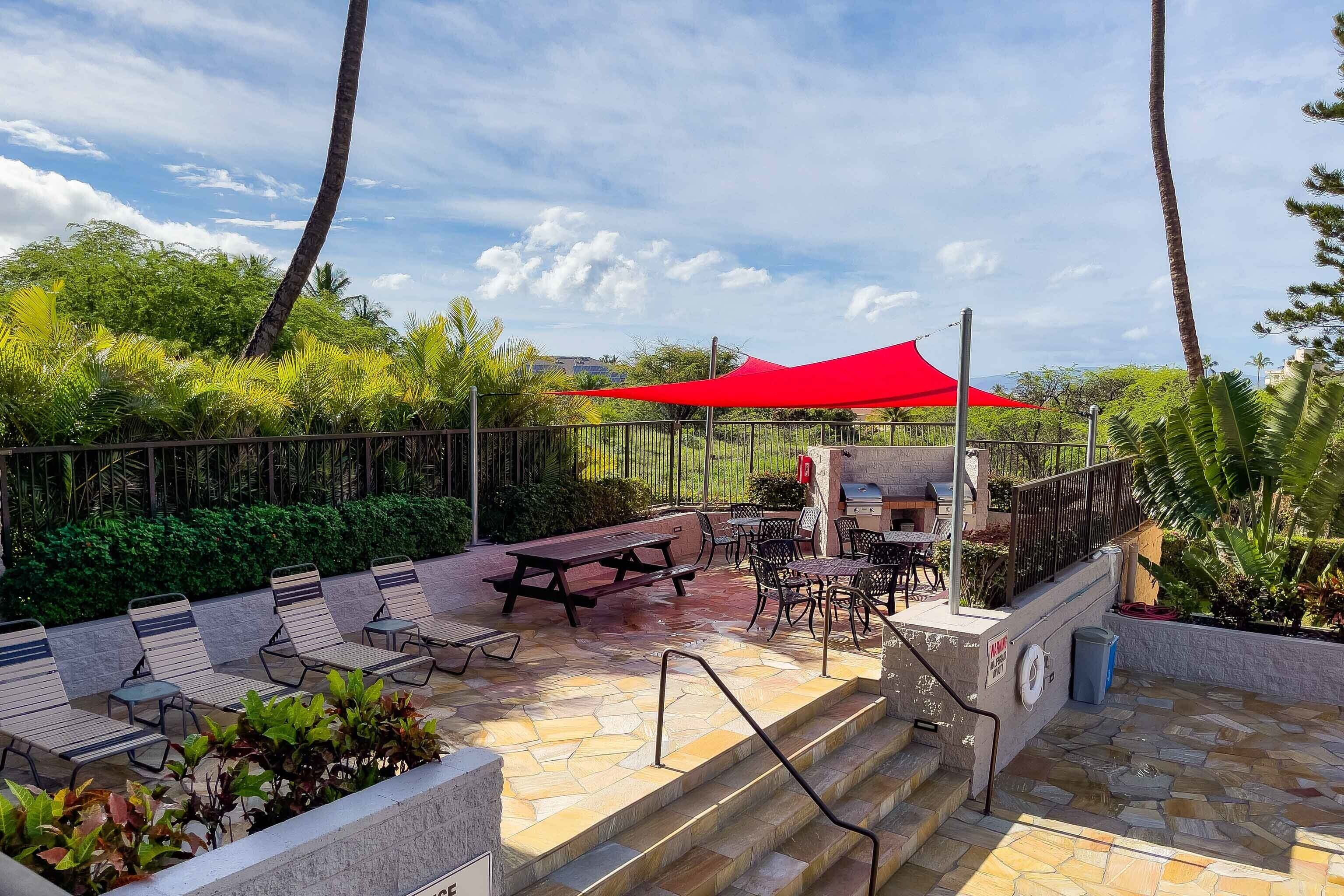 An outdoor patio with stone flooring, picnic tables, and chairs, surrounded by lush greenery and a red canopy.