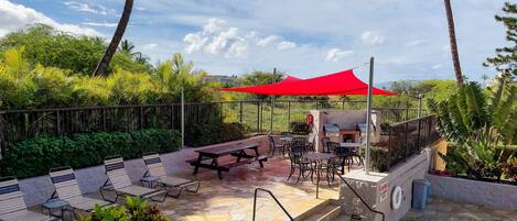An outdoor patio with stone flooring, picnic tables, and chairs, surrounded by lush greenery and a red canopy.