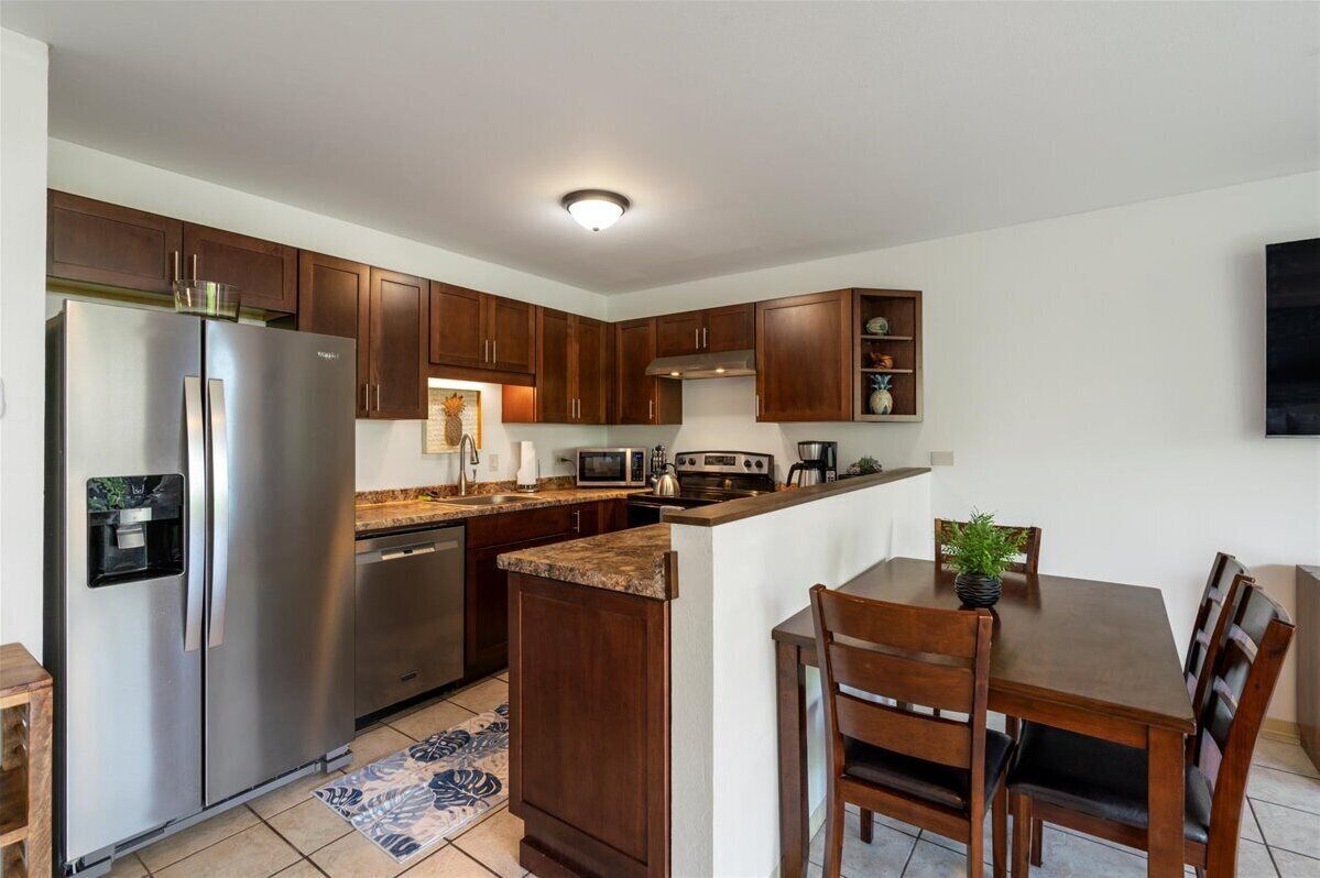 A modern kitchen with dark wooden cabinets, stainless steel appliances, a built-in microwave, and a dining area with wooden chairs and a small potted plant.