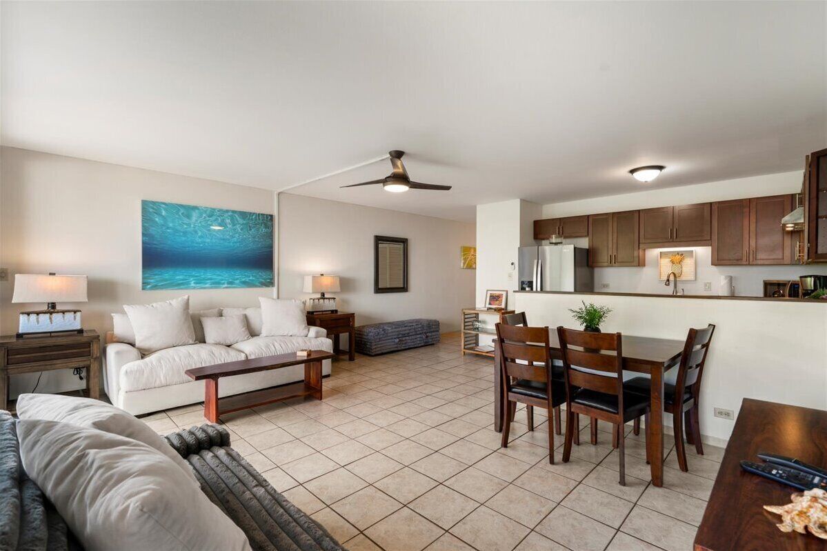Un vaste espace de vie comprenant un canapé blanc, une table basse et une table à manger avec des chaises. On y trouve également un ventilateur de plafond, un coin cuisine avec des meubles en bois et un grand tableau représentant une scène sous-marine accroché au mur.