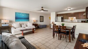 A spacious living area with a white sofa, a coffee table, and a dining table with chairs. There is a ceiling fan, a kitchen area with wooden cabinets, and a large painting of an underwater scene on the wall.