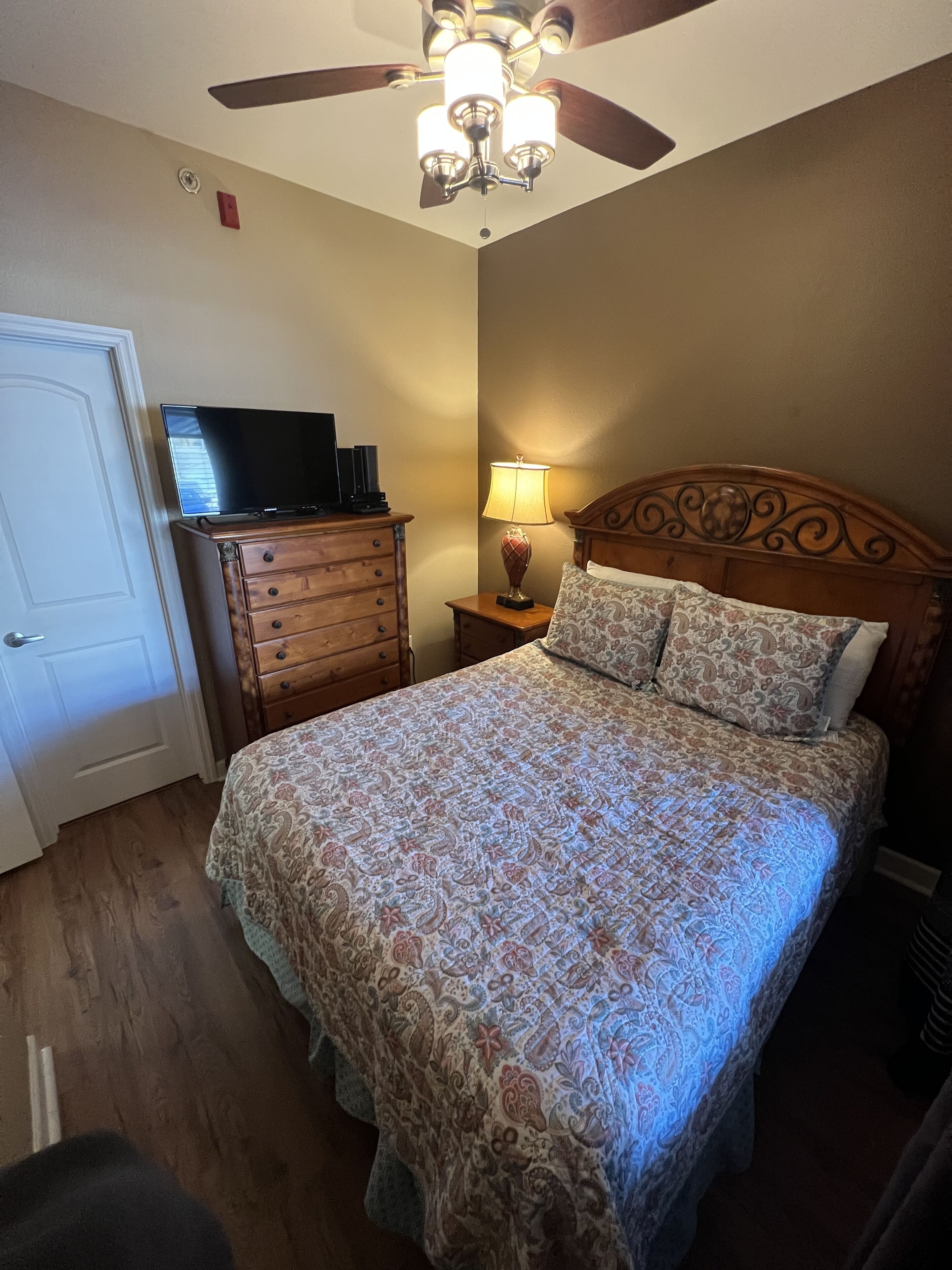 Quarto com cama, cômoda, televisão, ventilador de teto e mesa de cabeceira com abajur.